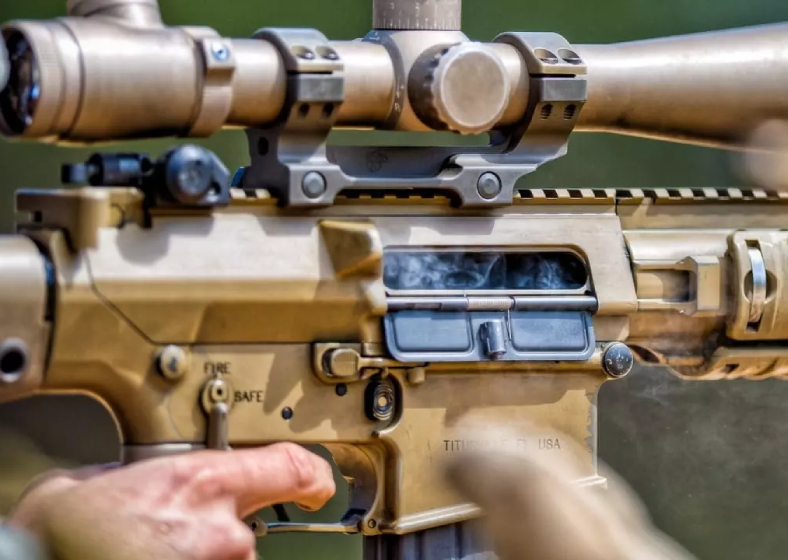 Close-up of a person's hand near a tan sniper rifle scope and receiver.